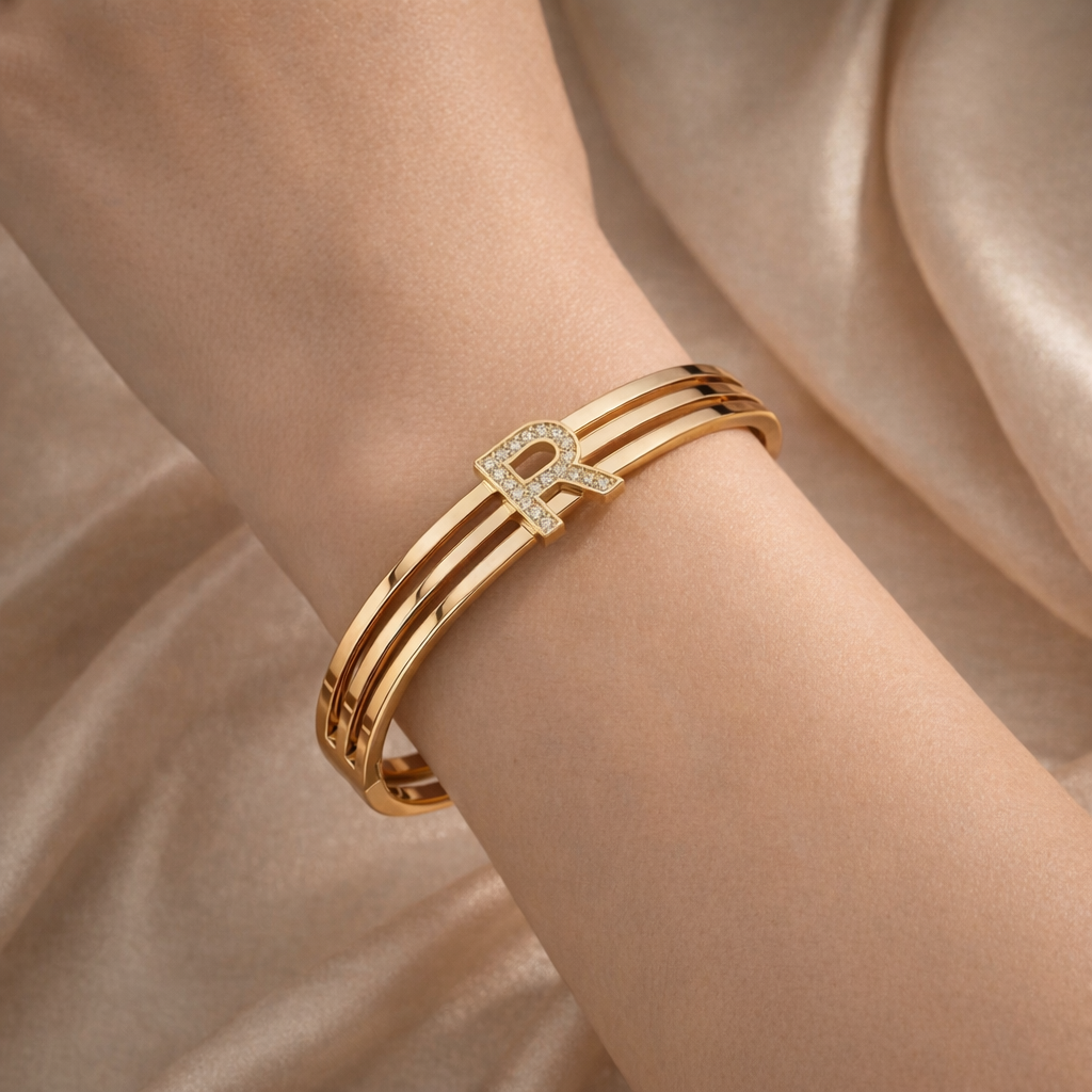 Gold bracelet with a diamond 'R' on a person's wrist against a beige fabric background