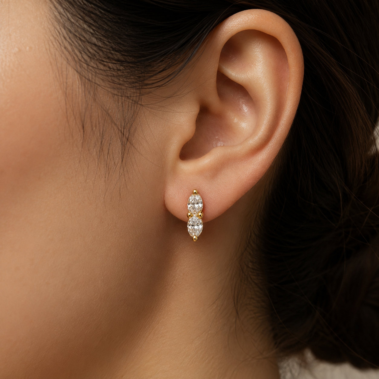 A close-up of a person's ear wearing a small gold huggie hoop earring with three marquise-cut clear stones.