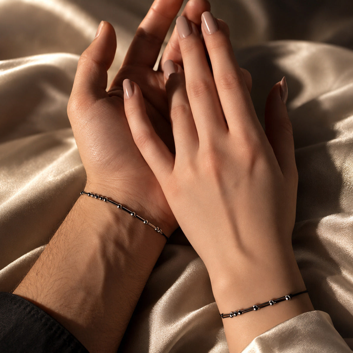 Two hands resting on draped champagne satin fabric, each wearing a delicate black beaded morse code bracelet.