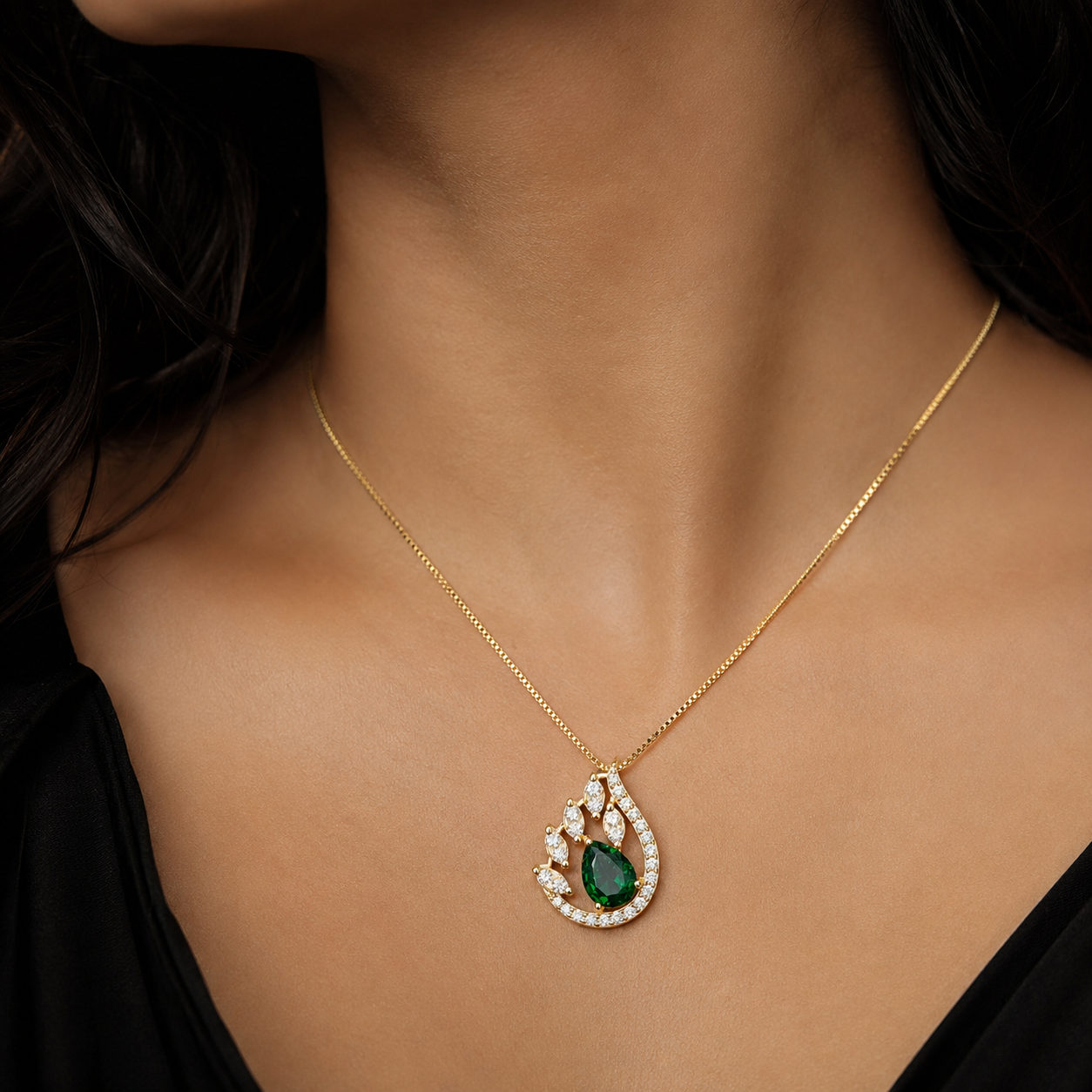 model's neck wearing a delicate gold chain necklace with a teardrop-shaped pendant. The pendant features a central green emerald-cut stone surrounded by small clear stones, all set in a gold frame.