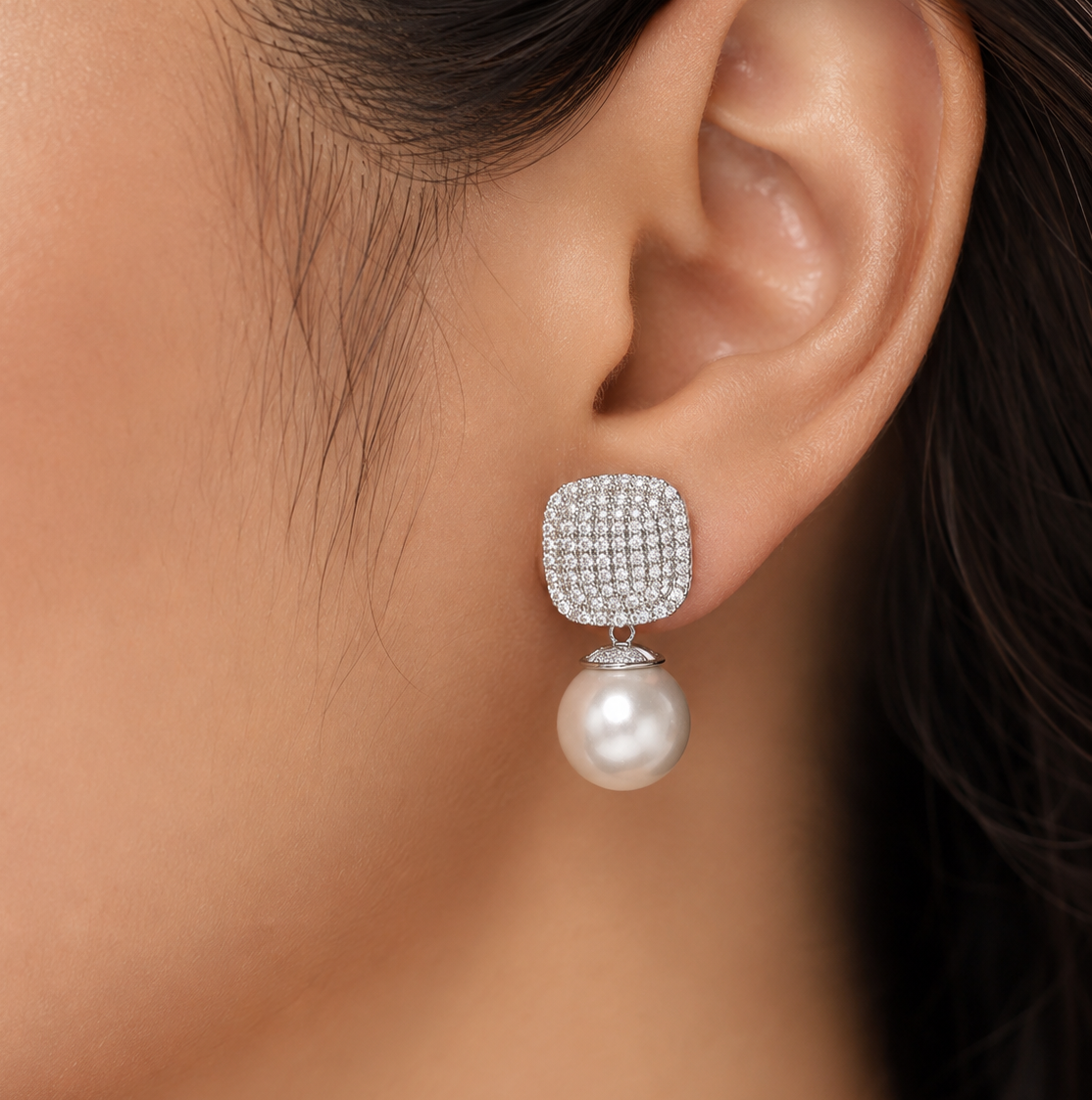woman's ear wearing a silver-toned square stud earring paved with clear diamonds and dangling a single white pearl